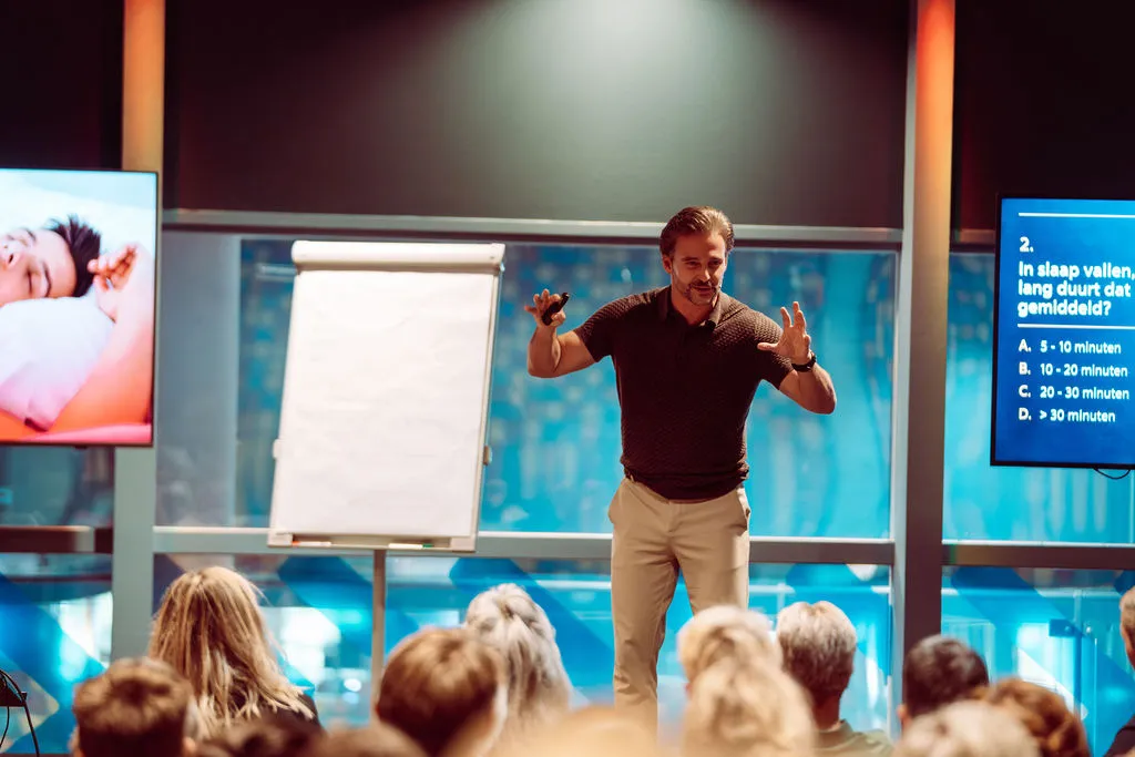 Dr. Merijn van de Laar staat voor een zaal met een flip-over, naast een scherm met de quizvraag hoe lang inslapen gemiddeld duurt