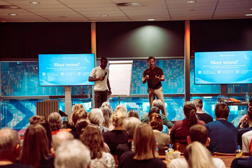 Dr. Merijn van de Laar op een podium naast een presentator, voor een volle zaal, met op de schermen Meer weten? deslaapwetenschapper.nl