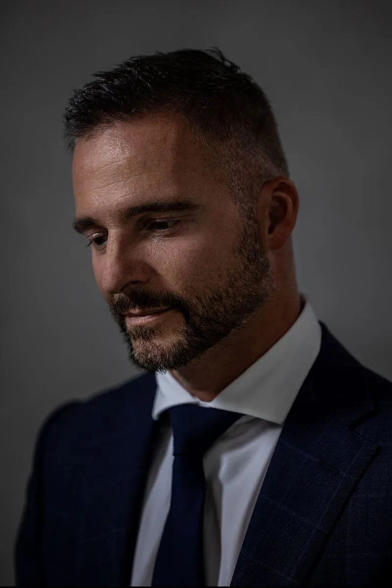 Dr. Merijn van de Laar in een donkerblauw pak met stropdas kijkt naar beneden, tegen een donkergrijze achtergrond