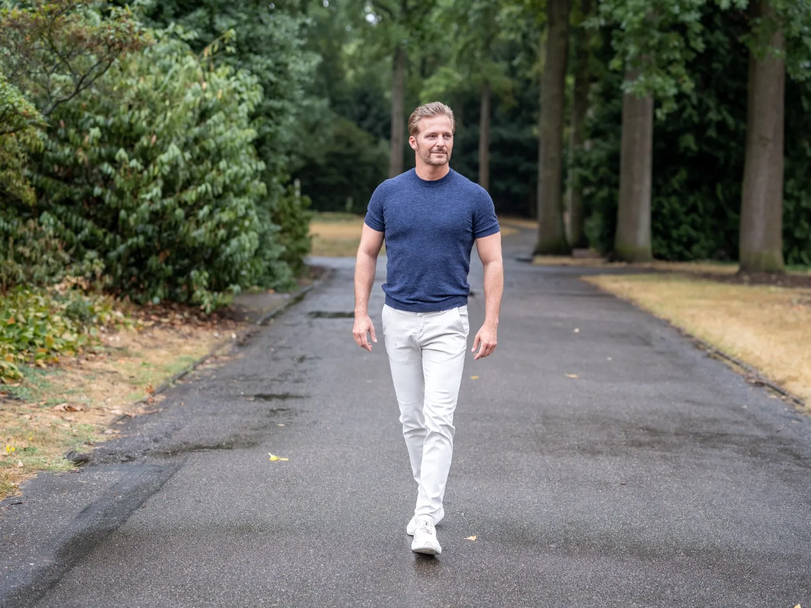 Dr. Merijn van de Laar wandelt over een laan met hoge bomen, in een blauw shirt en een witte broek